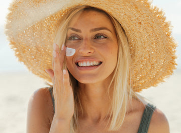 Woman applying sunscreen on her face with a straw hat on a beach.